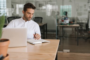 Odaklanmış genç işadamı bir dizüstü bilgisayar ile online çalışma ve modern bir ofiste masasında tek başına otururken notlar yazma