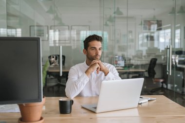 Odaklanmış genç işadamı modern bir ofiste masasında bir dizüstü bilgisayar ile online çalışırken düşünce derin görünümlü