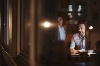 İki genç işadamı, gece geç saatlerde bir masada birlikte çalışırken gülümseyen ofis binası pencerelerini şehir yansıtan