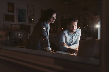 İki genç işadamı, akşam ları ofisi yansıtan pencerelerin arkasındaki masada birlikte çalışırken gülümseyerek