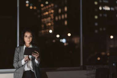 Odaklanmış genç iş kadını gece geç saatlerde bir ofiste tek başına arka planda şehir ışıkları ile bir dijital tablet ile çalışan ayakta
