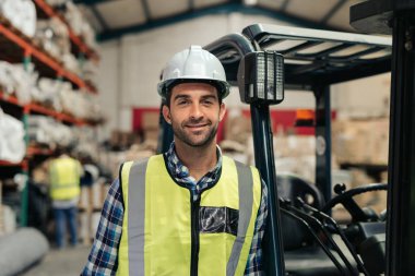 Bir halı deposunun zemininde dururken sert hat ve yelek giyen gülümseyen forklift sürücüsünün portresi