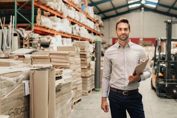 Portrait of a manager holding a clipboard while standing on the warehouse floor with stacks of carpets and textiles on shelves in the background
