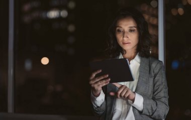 Odaklanmış genç iş kadını arka planda şehir ışıkları ile gece geç saatlerde bir dijital tablet ile çalışan bir ofiste tek başına ayakta