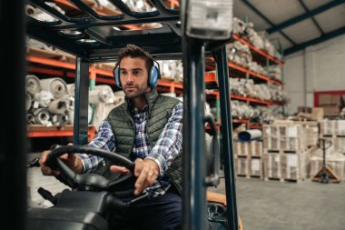 Forklift sürücüsü büyük bir halı deposunun zemin etrafında stok hareket ederken koruyucu kulak muffs giyiyor