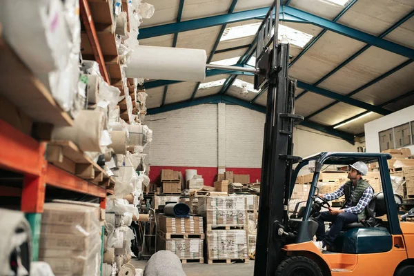 Forklift driver carefully transporting a carpet role from a shelf while working on the floor of a large warehouse