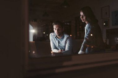 İki genç iş arkadaşı, gece geç saatlerde bir masada birlikte bir dizüstü bilgisayar kullanarak ofis binası pencerearkasında şehir yansıtan