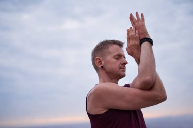 Fit adam alacakaranlıkta dramatik bir gökyüzüne karşı okyanus kenarında tek başına yoga yaparken kartal poz yapıyor