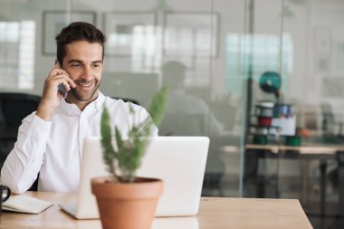 Gülümseyen genç işadamı bir dizüstü bilgisayar üzerinde çalışan modern bir ofiste masasında otururken cep telefonu yla konuşuyor