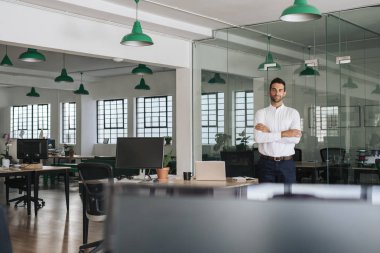 Kolları çapraz büyük bir modern ofiste tek başına dururken gülümseyen kendine güvenen genç bir işadamı portresi