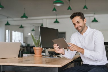 Gülümseyen genç işadamı büyük bir modern ofiste masasında otururken dijital bir tablet ile online çalışan