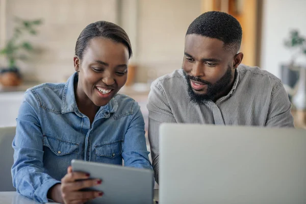 Smiling young African American couple sitting together at their dining table using a laptop and a digital tablet