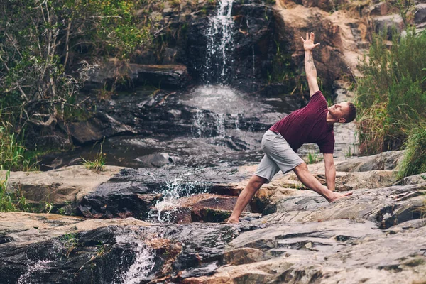 Fit man doing the triangle pose while practicing yoga on some rocks by a waterfall in a forest