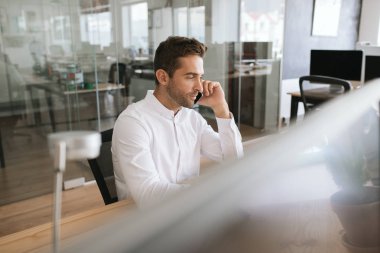 Odaklanmış genç işadamı modern bir ofiste masasında tek başına bir cep telefonu bir müşteri ile konuşurken oturuyor