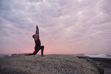 Fit adam gün batımında dramatik bir gökyüzüne karşı okyanus tarafından bazı kayalar üzerinde yoga yaparken diz poz cresent lunge yapıyor