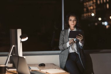 Ofis masasının yanında duran genç iş kadını, gece geç saatlerde arka planda şehir ışıkları olan dijital bir tabletle çevrimiçi çalışıyor