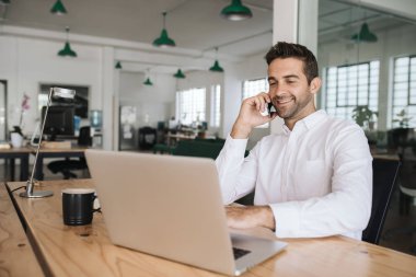 Gülümseyen genç işadamı modern bir ofiste masasında oturan bir dizüstü bilgisayar üzerinde çalışıyor ve onun cep telefonu konuşuyor