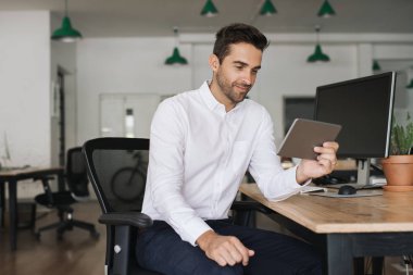 Genç işadamı dijital tablet ile online çalışan büyük bir modern ofiste masasında otururken gülümseyen