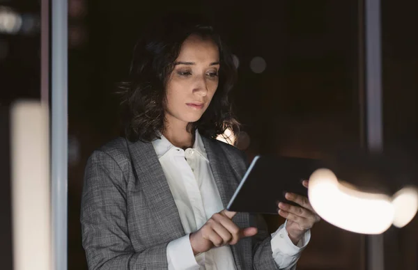 Young businesswoman working overtime while standing in a dark office working online with a digital tablet