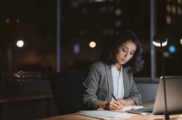 夜遅くにオフィスデスクで残業しながら ノートパソコンを使ってメモを書く若いビジネスウーマン — ストック写真