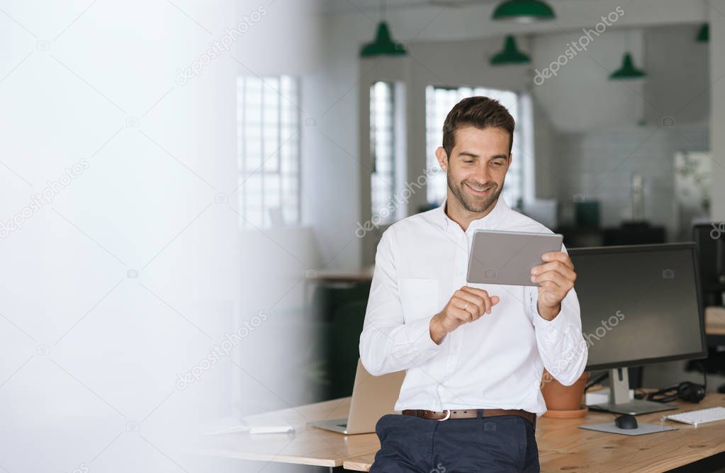 Smiling young businessman leaning against a desk in a large modern office working online with a digital tablet