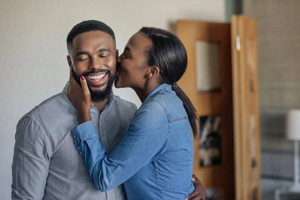 若いアフリカ系アメリカ人の女性は 彼女の笑顔の夫に自宅のリビングルームで頬にキスを与える — ストック写真