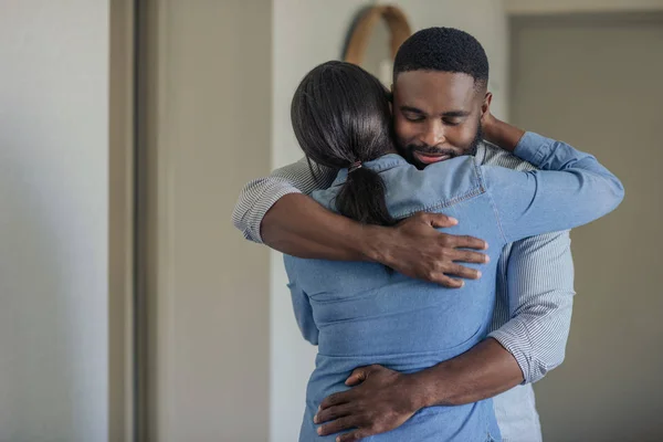 目を閉じた若いアフリカ系アメリカ人男性が 自宅の居間で一緒に立っている間 妻を抱きしめている — ストック写真