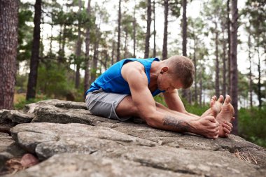 Bir çam ormanında tek başına yoga yaparken arka streç poz yapıyor kayalık bir çıkıntı üzerinde oturan adam