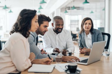 Modern bir ofisin yönetim kurulu toplantısında bir dizüstü bilgisayar üzerinde birlikte çalışan gülümseyen işadamlarından oluşan farklı bir grup