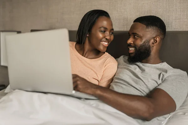 微笑的年轻非洲裔美国夫妇一起在笔记本电脑上观看视频 而躺在睡衣在他们的床上在早上 — 图库照片