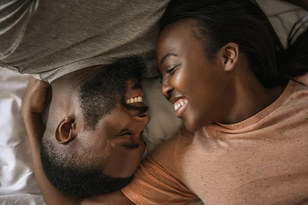 若いアフリカ系アメリカ人のカップルは 朝一緒にベッドに向かって横たわっている間 愛情を込めてお互いに微笑み合う — ストック写真