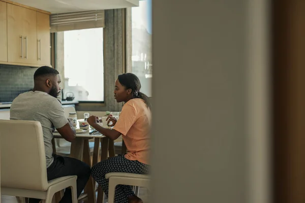 Young African American couple talking and eating breakfast together at their kitchen dining table in the morning