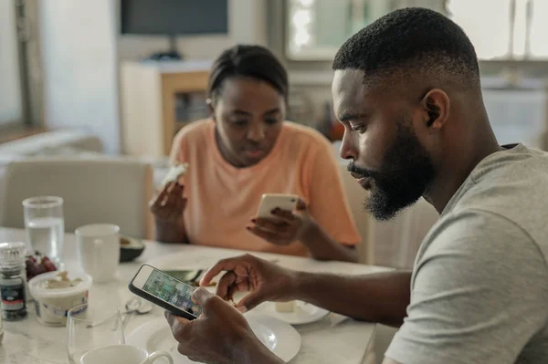 Young African American couple distracted by their cellphones while eating breakfast together at home in the morning