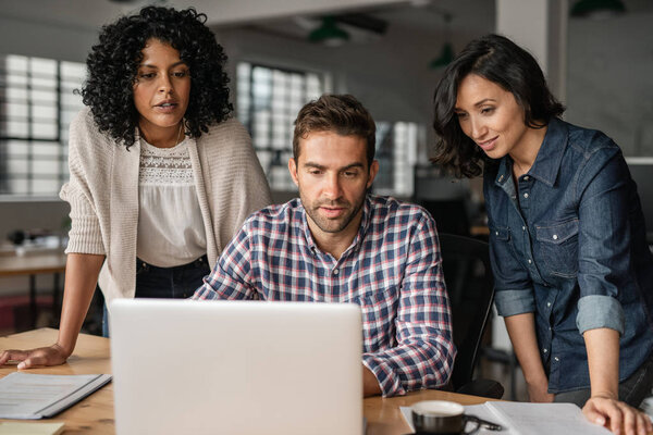 Трое молодых дизайнеров, работающих в сети вместе над ноутбуком за столом коллег в их стартапе
