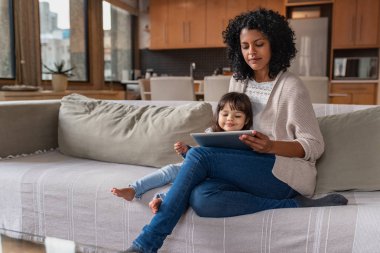 Sevimli küçük kız ve annesi evde oturma odası kanepede birlikte otururken bir dijital tablet kullanarak