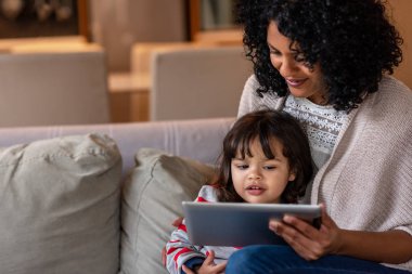 Gülümseyen anne ve onun sevimli küçük kızı birlikte bir dijital tablet üzerinde bir şey izlerken oturma odasında kanepede oturan