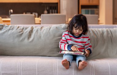 Sevimli küçük kız bir dijital tablet kullanarak evde oturma odasında bir kanepede tek başına oturan
