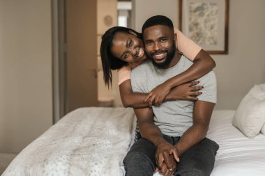 Sabahları yataklarında pijamalarıyla otururken kocasına arkadan sarılan gülümseyen genç bir Afro-Amerikan kadının portresi