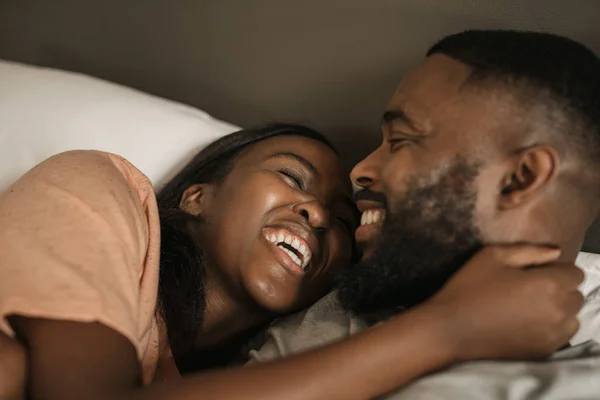 Affectionate young African American couple laughing and hugging each other while lying in bed together in the morning