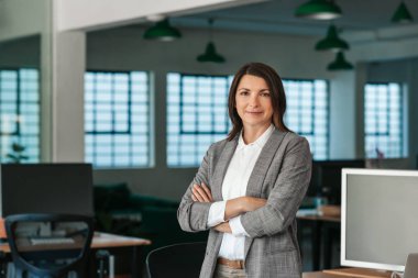 Öğleden sonra büyük, modern bir ofiste kollarını kavuşturarak duran gülümseyen bir iş kadınının portresi.