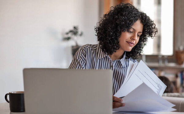 Smiling young female entrepreneur going over paperwork while working on a laptop at her dining room table at home