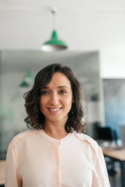 Öğleden sonra büyük, modern bir ofiste tek başına dururken gülümseyen genç bir iş kadınının portresi.