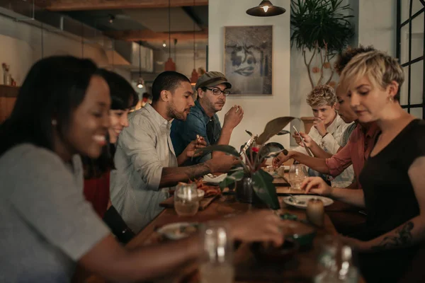 不同的一群年轻朋友一边吃着食物 一边笑着 晚上一起坐在餐厅的餐桌前 — 图库照片