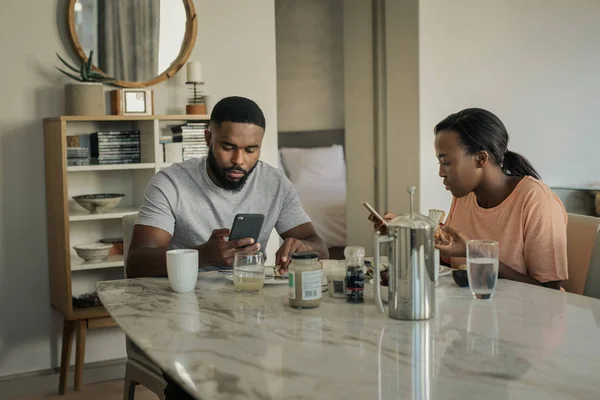 Young African American couple distracted by their cellphones while having breakfast together at home in the morning