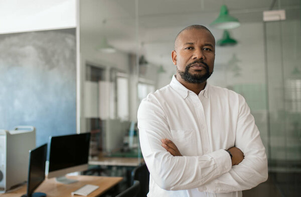 Portrait of a confident mature African American businessman standing with his arms crossed in a large modern office in the late afternoon