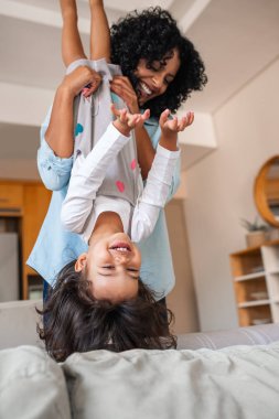 Gülümseyen anne, evde birlikte oynarken küçük kızını kanepelerinde baş aşağı tutarak gülüyor.