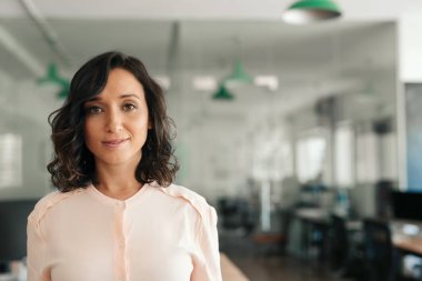 Öğleden sonra büyük, modern bir ofiste tek başına dururken gülümseyen bir iş kadını portresi.