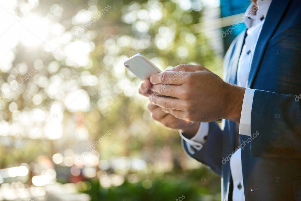 Closeup of a businessman sending a text message on his cellphone while standing outside in the city on a sunny afternoon