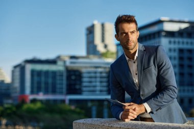 Odaklanmış genç işadamı bir ofis terasına yaslanıyor ve elinde dijital tablet tutuyor.