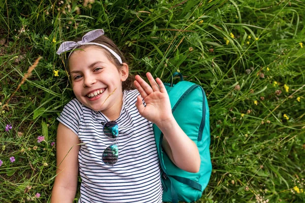 Cute Little Girl Uśmiecha Się Macha Cześć Leżąc Plecakiem Trawie — Zdjęcie stockowe